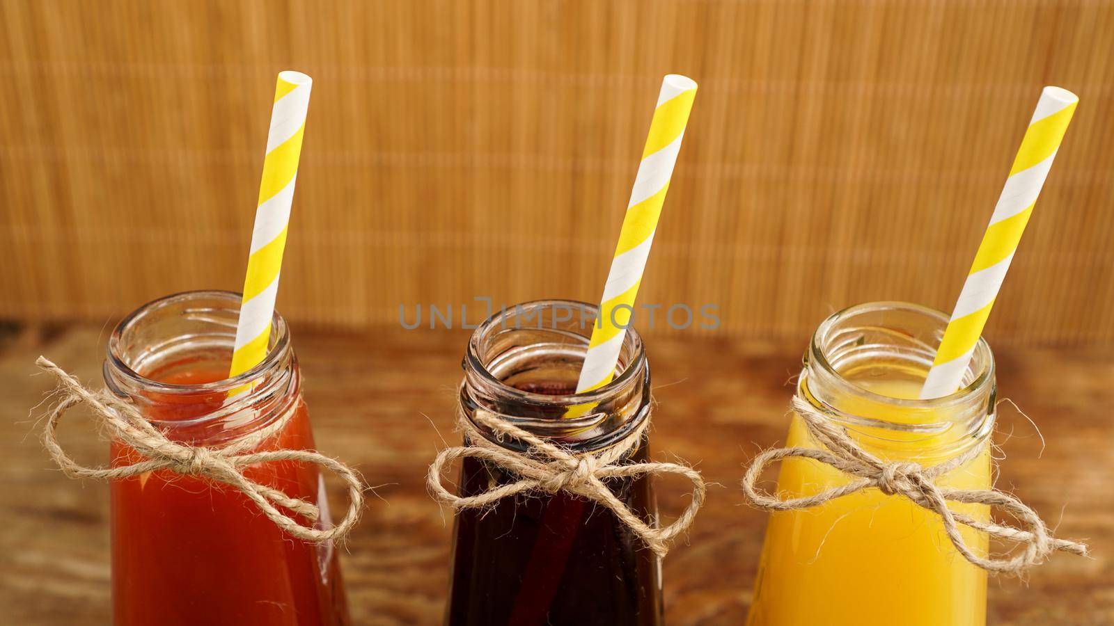 Three multicolored drinks in the bottles on a wooden bamboo background. Bottled paper straws. The bottle is tied with twine. Homemade drink