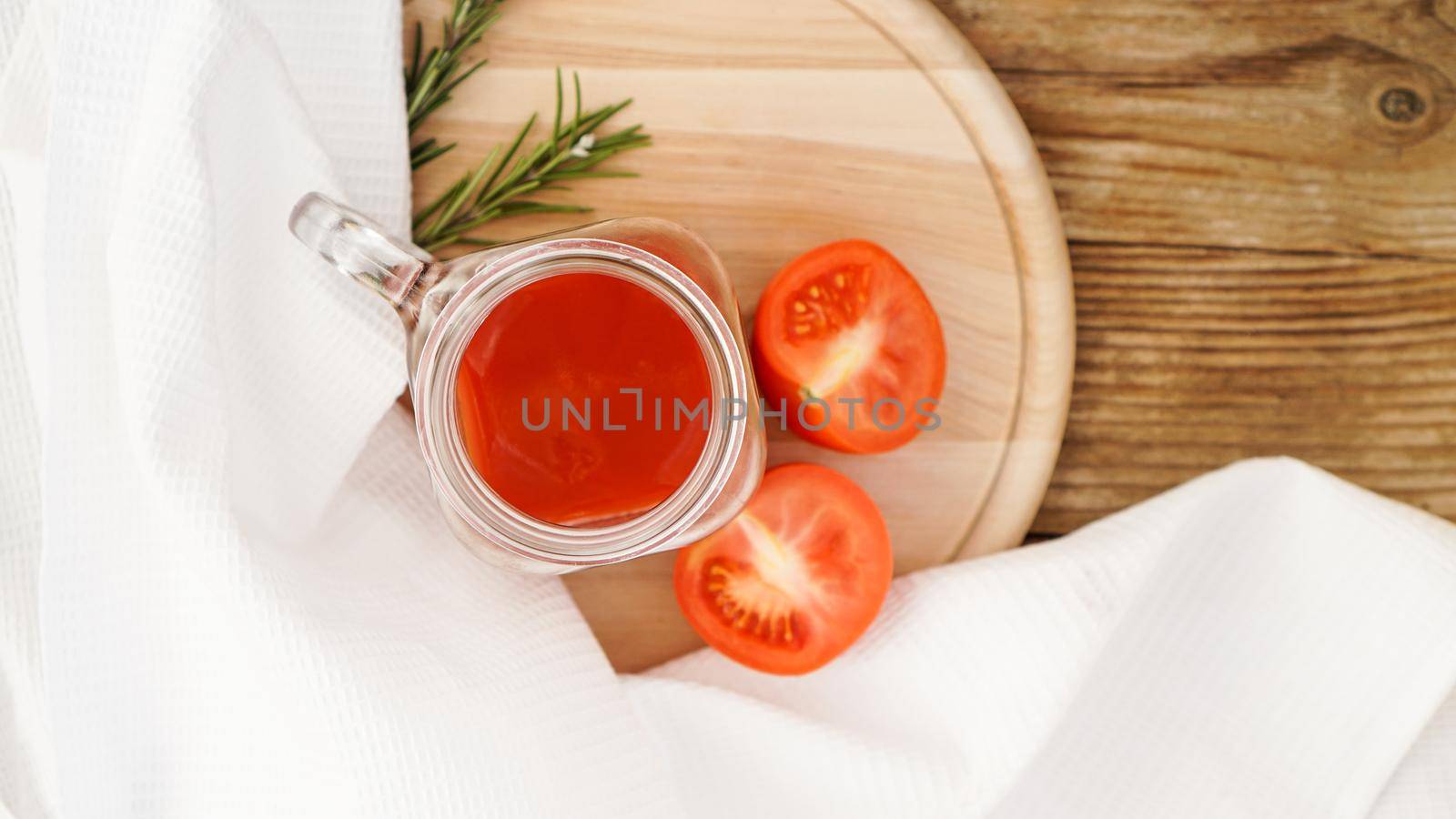 Tomato juice in glass jar, fresh tomatoes and sprigs of rosemary on wooden cutting board and white towel - Top view