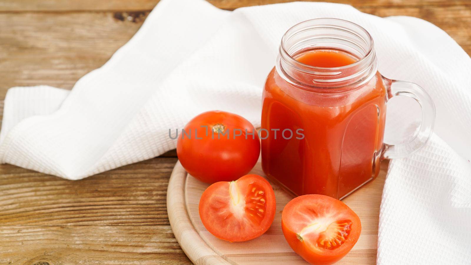 Tomato juice in glass jar and fresh tomatoes on wooden cutting board and white towel