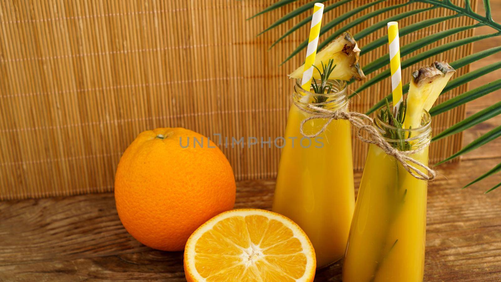 Two bottles of tropical juice with paper straws. Oranges, pineapple and rosemary for decoration. Wooden background.