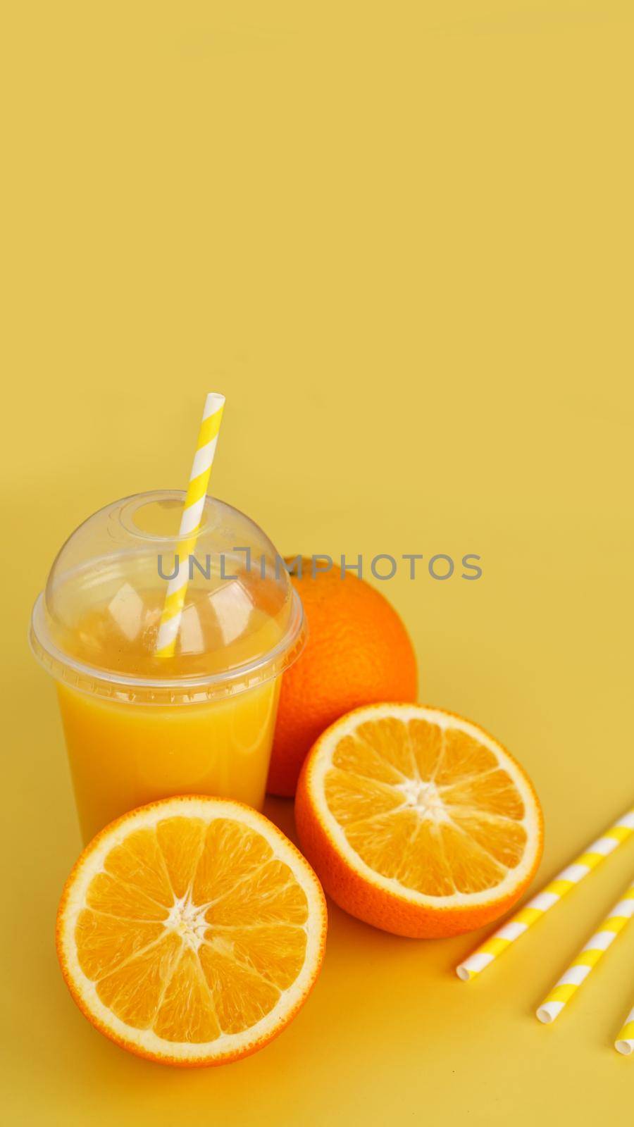 Orange juice in fast food closed cup with tube on yellow background. Sliced orange and yellow paper straws for a drink. Vertical photo