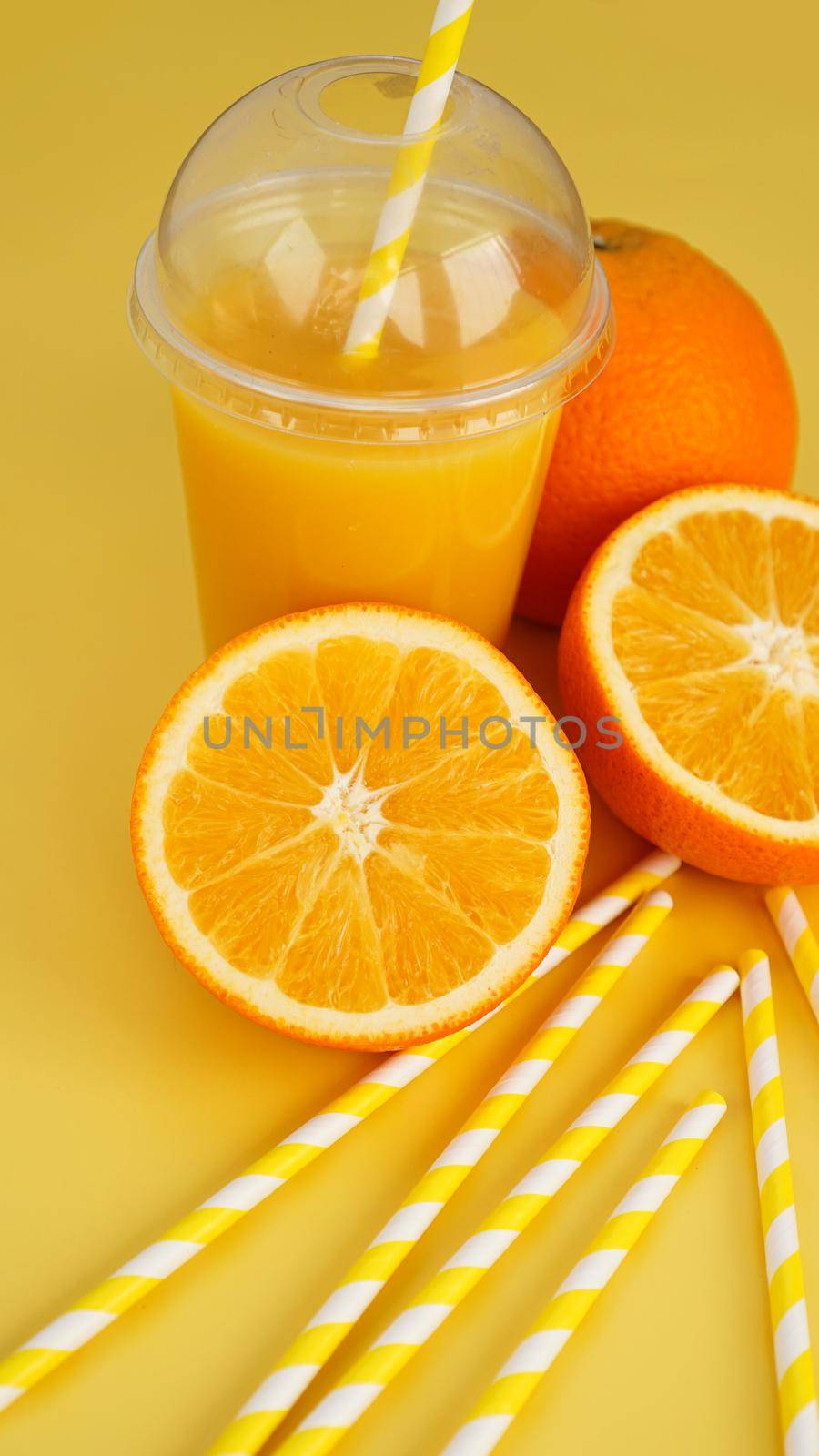 Orange juice in fast food closed cup with tube on yellow background. Sliced orange and yellow paper straws for a drink. Vertical photo