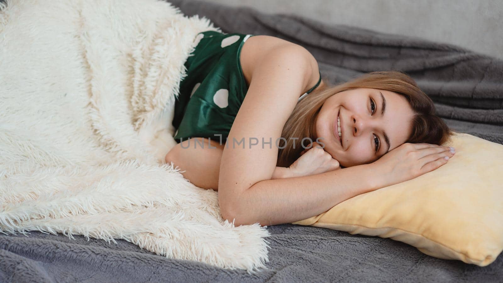 Beautiful smiling blonde in green pajamas on a yellow pillow on a sofa under a white blanket. Good happy morning concept