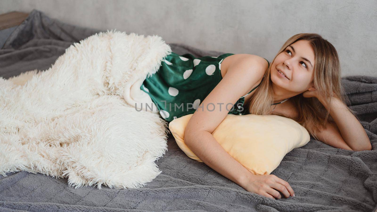 A beautiful girl lies on sofa under a white blanket. She just woke up and thinks about plans for the day. Beautiful smiling blonde in green pajamas.