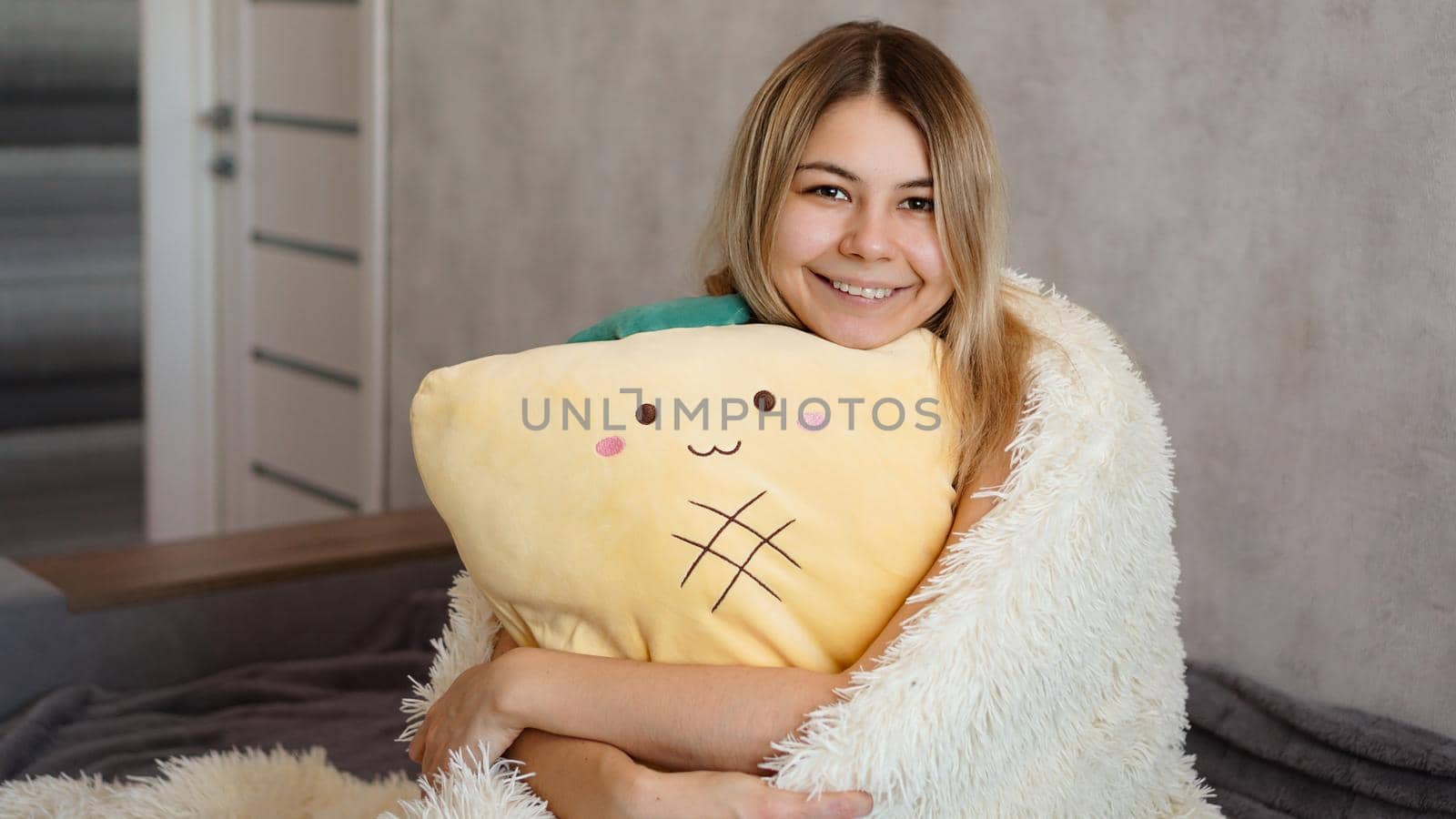 Happy blonde under a white blanket in the morning hugs a yellow pillow. Happy morning concept