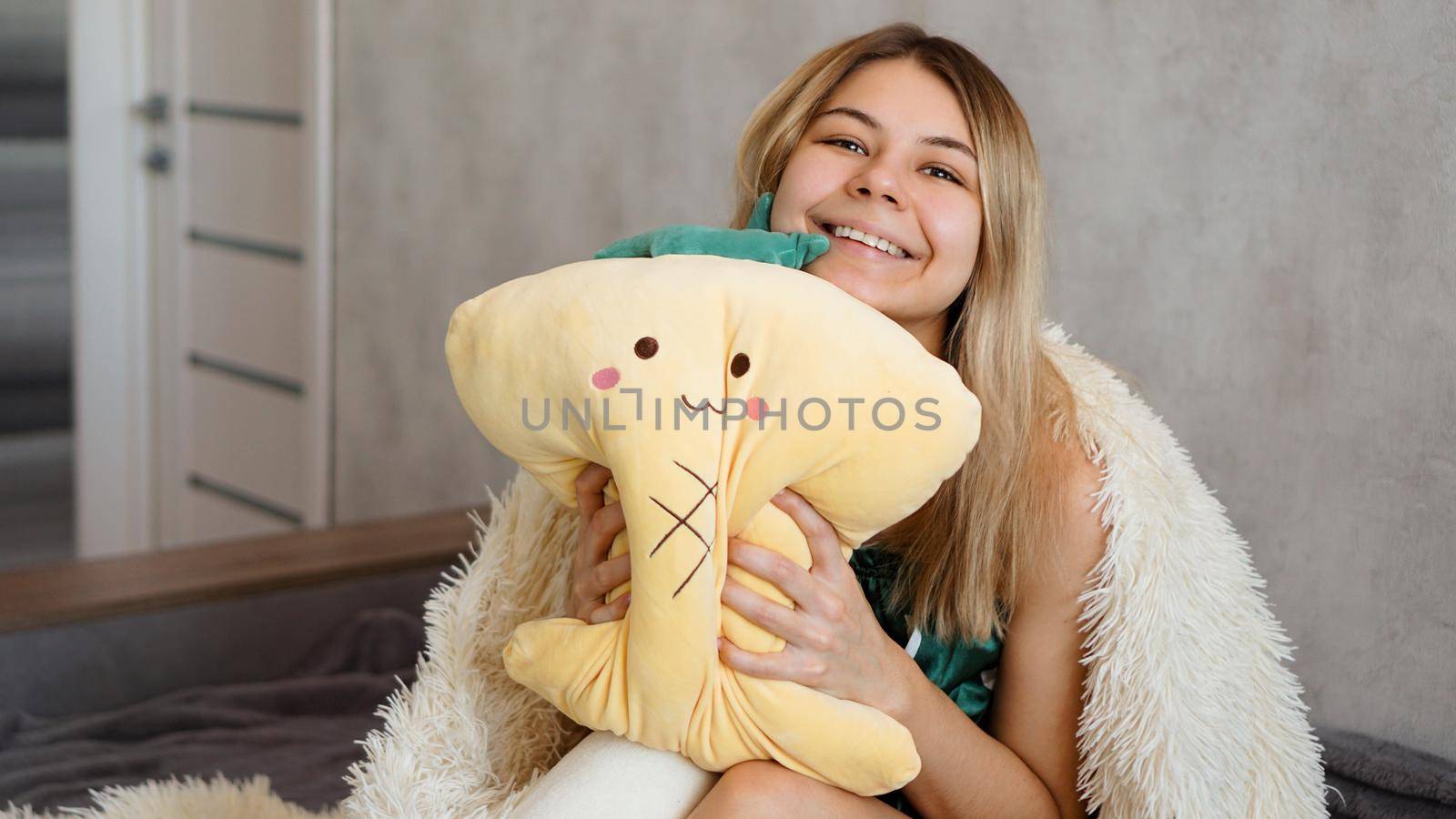 Happy blonde under a white blanket in the morning hugs a yellow pillow. Happy morning concept