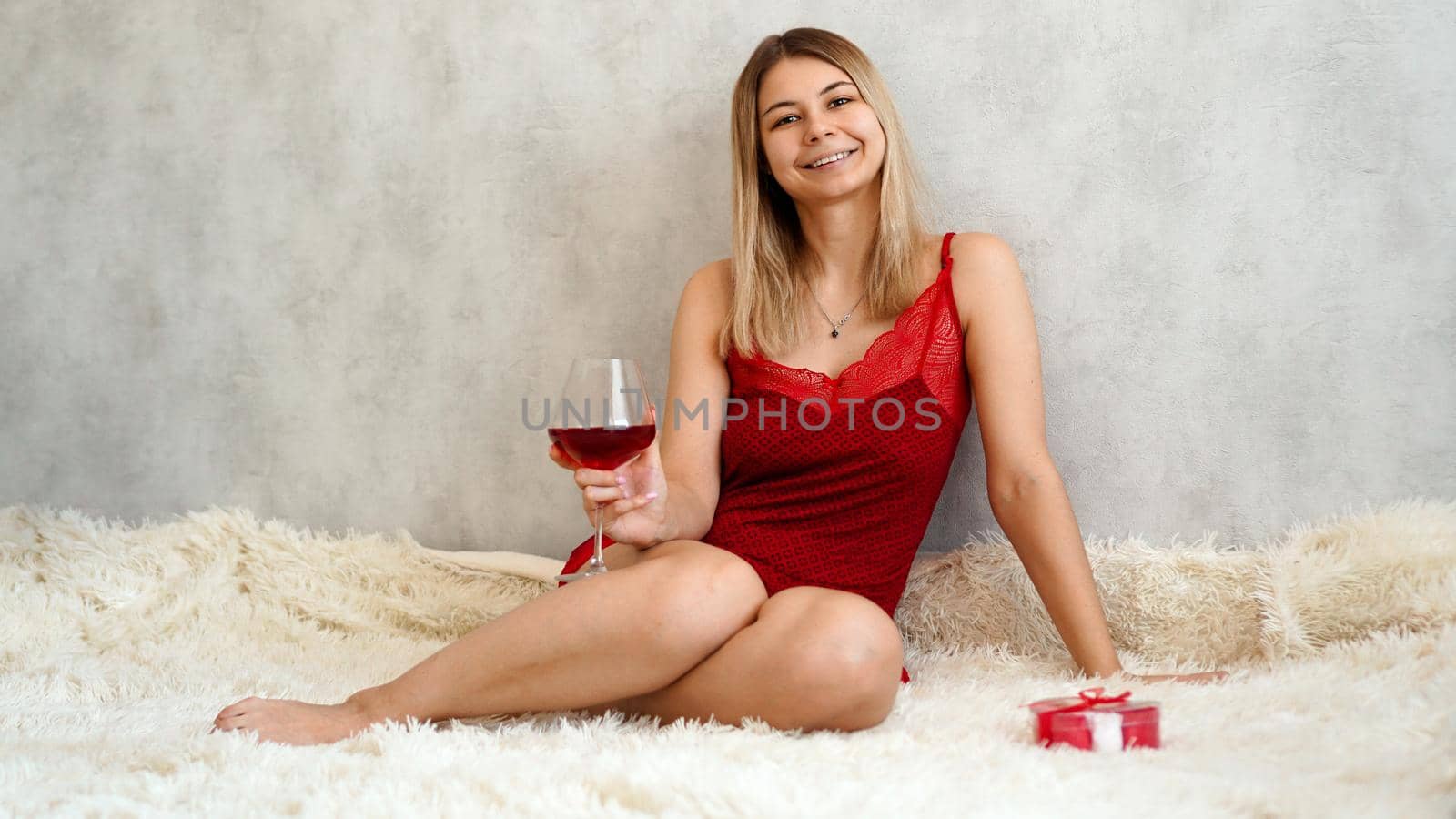 A beautiful young woman is sitting in red underwear on a white plaid with wine in her hand. Valentines day morning