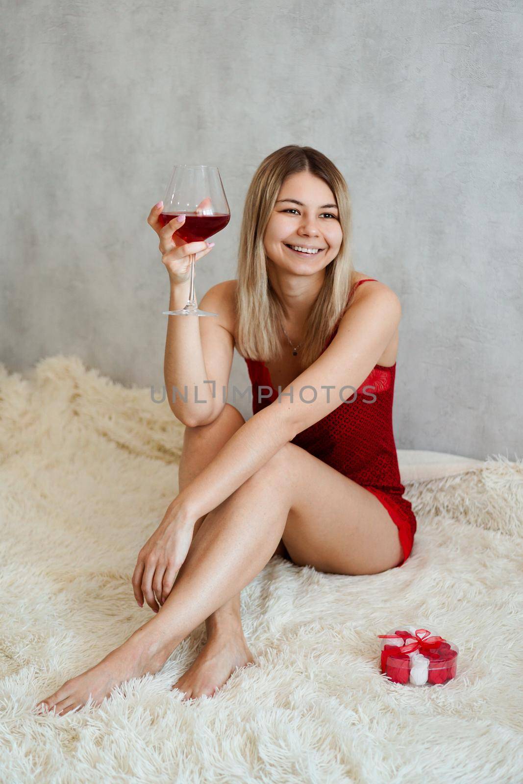 A beautiful young woman with smile is sitting in red underwear on a white plaid with wine in her hand. Valentines day morning. Vertically photo