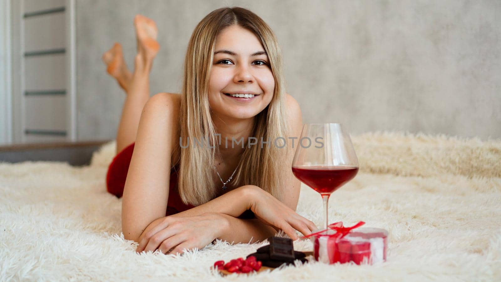 A beautiful smiling blonde lies in bed. Valentines Day Morning. A glass of wine, chocolate, sweets and a gift next to the girl. Happy morning in love