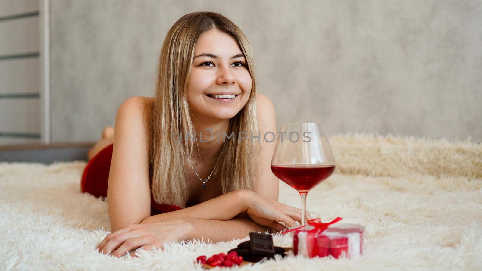 A beautiful smiling blonde lies in bed. Valentines Day Morning. A glass of wine, chocolate, sweets and a gift next to the girl. Happy morning in love