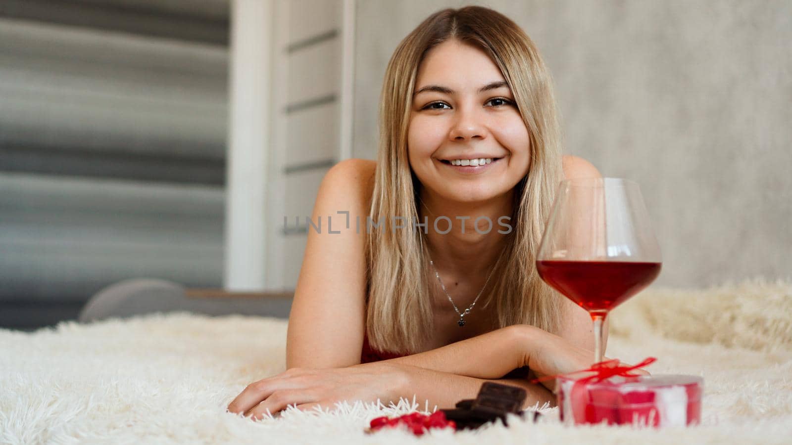 A beautiful smiling blonde lies in bed. Valentines Day Morning. A glass of wine, chocolate, sweets and a gift next to the girl. Happy morning in love