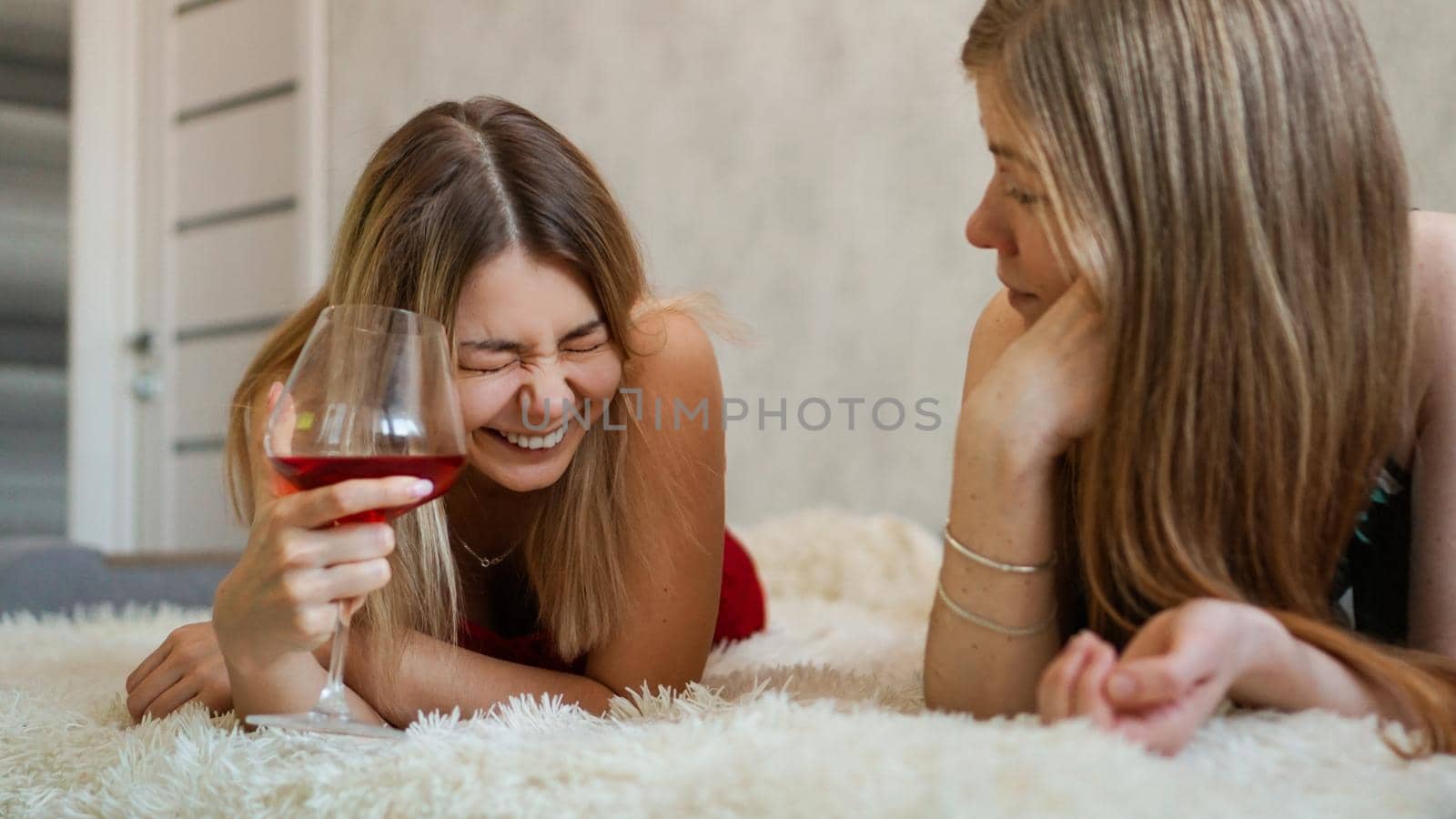 Two friends are lying on the couch and chatting. Beautiful blonde holding a glass of red wine in her hand