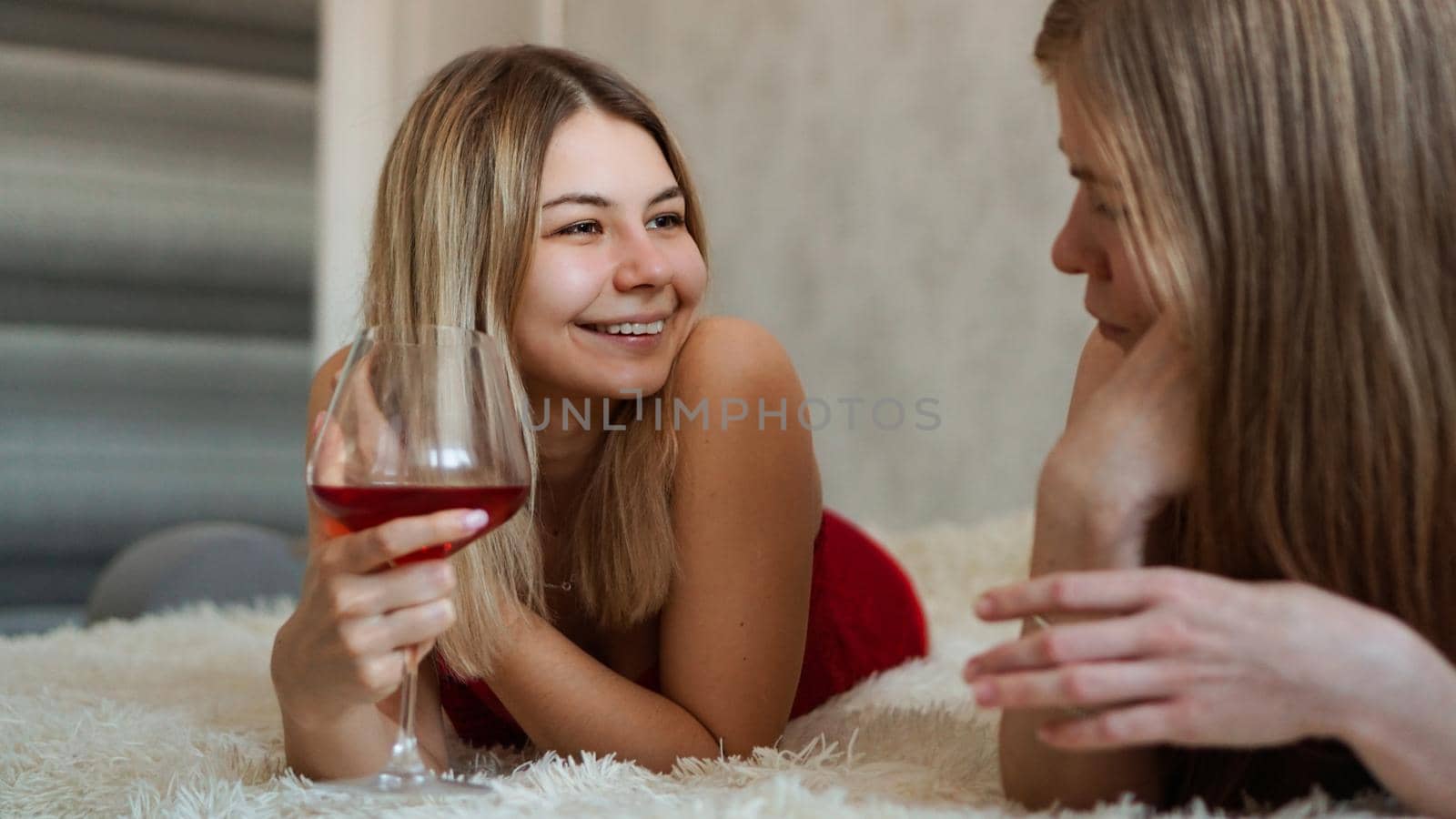 Two friends are lying on the couch and chatting. Beautiful blonde holding a glass of red wine in her hand