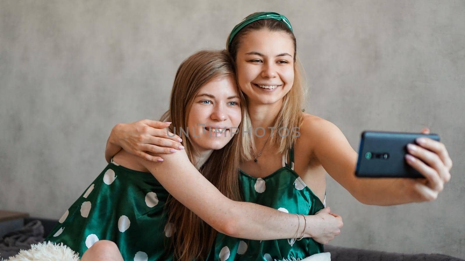 Two beautiful happy girlfriends hugging and taking selfies with smartphone at pajama party