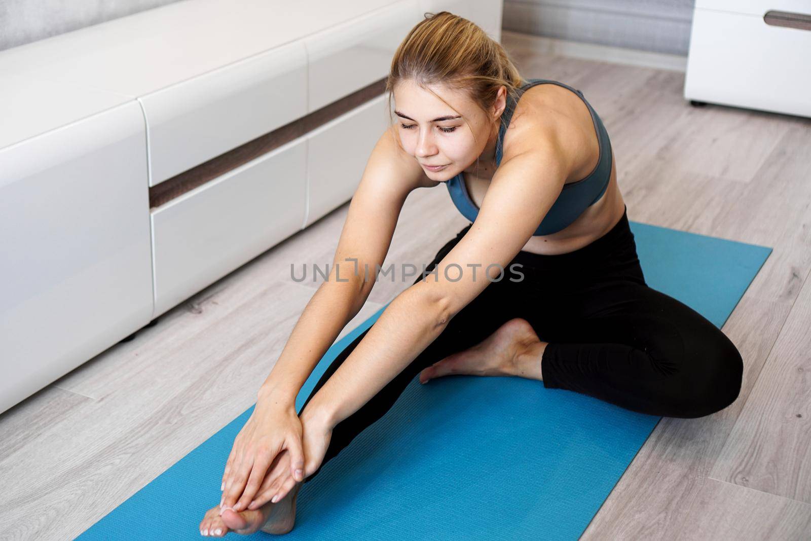 Sport, training and lifestyle concept - woman stretching leg on blue yoga mat at home in the living room