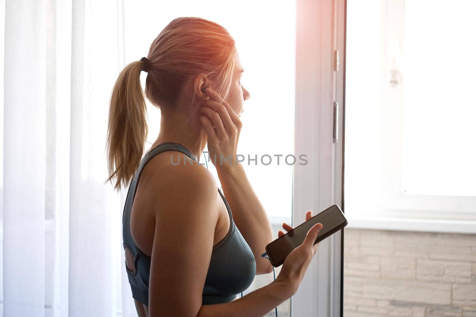 Young pretty sporty girl stands near the window and listens to music on headphones from smartphone. Sunny day