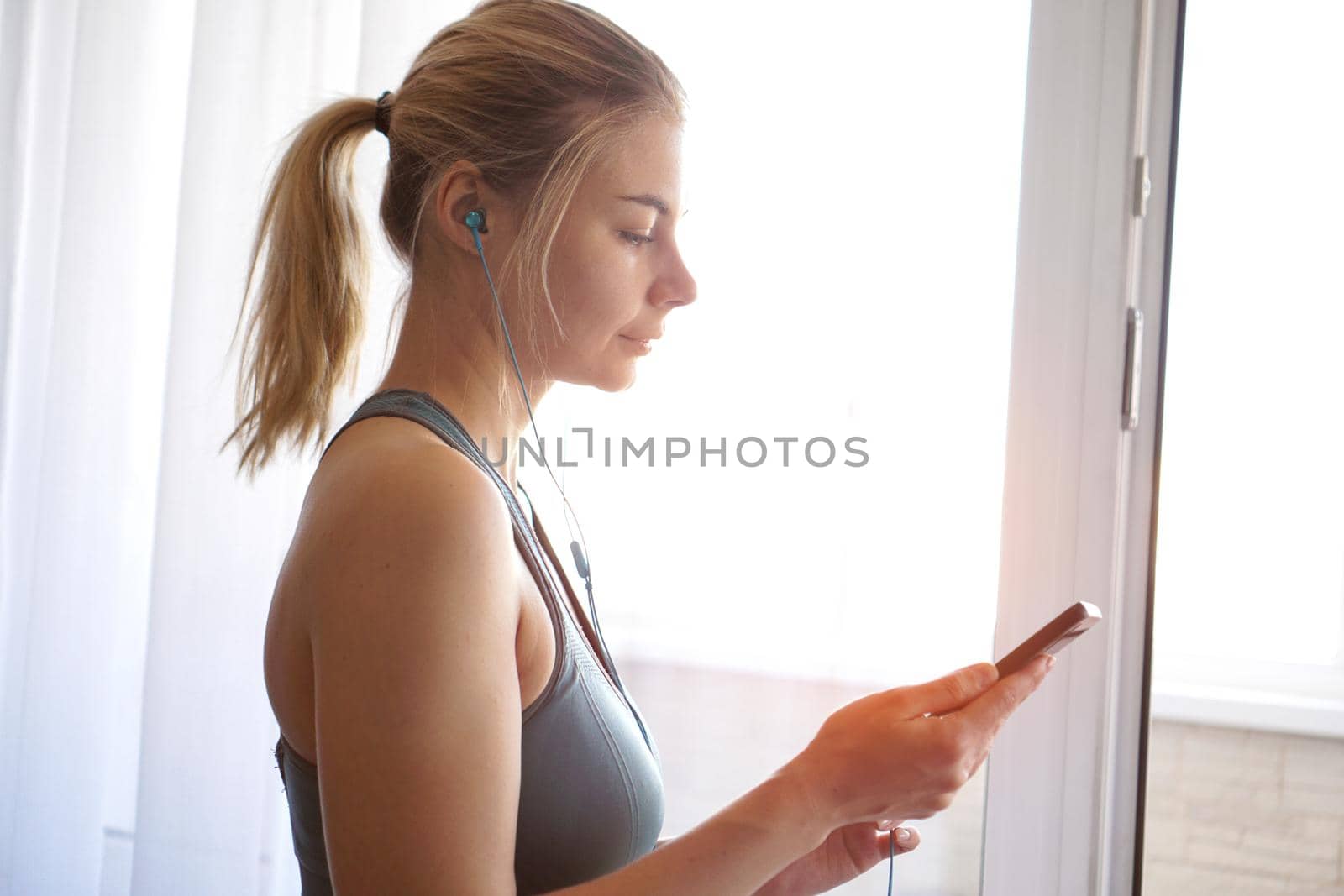 Young pretty sporty girl stands near the window and listens to music on headphones from smartphone. Sunny day