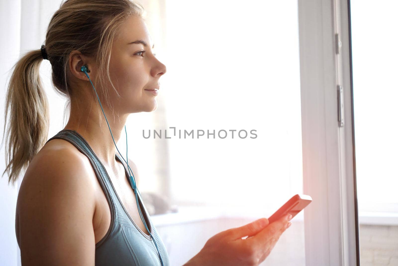 Happy young pretty sporty girl stands near the window and listens to music on headphones from smartphone