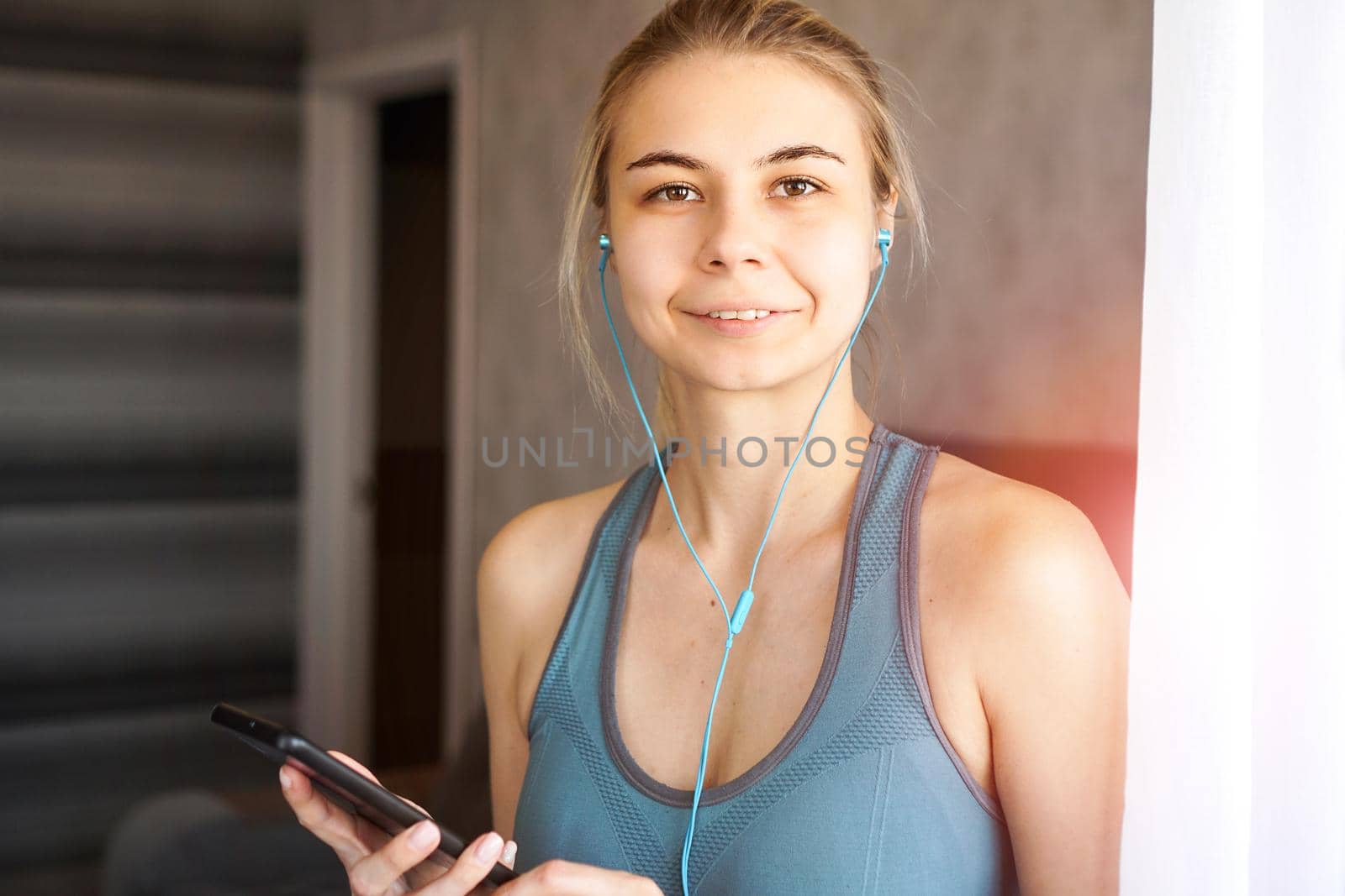 Young pretty sporty girl at home listens to music on headphones from smartphone. Sunny day