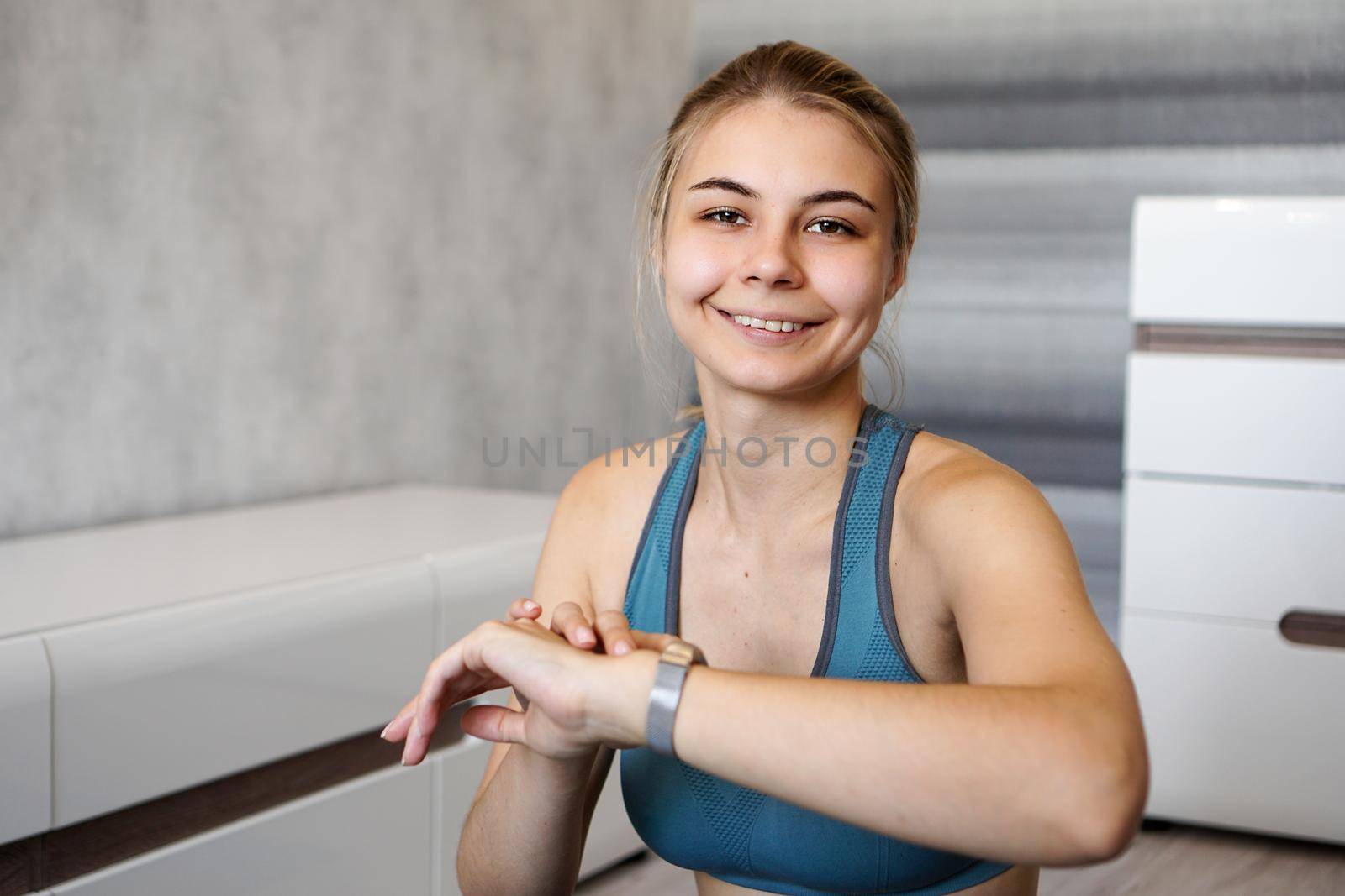 Portrait of young woman checking digital fitness tracker during self-training at home. She looks at the camera and smiles