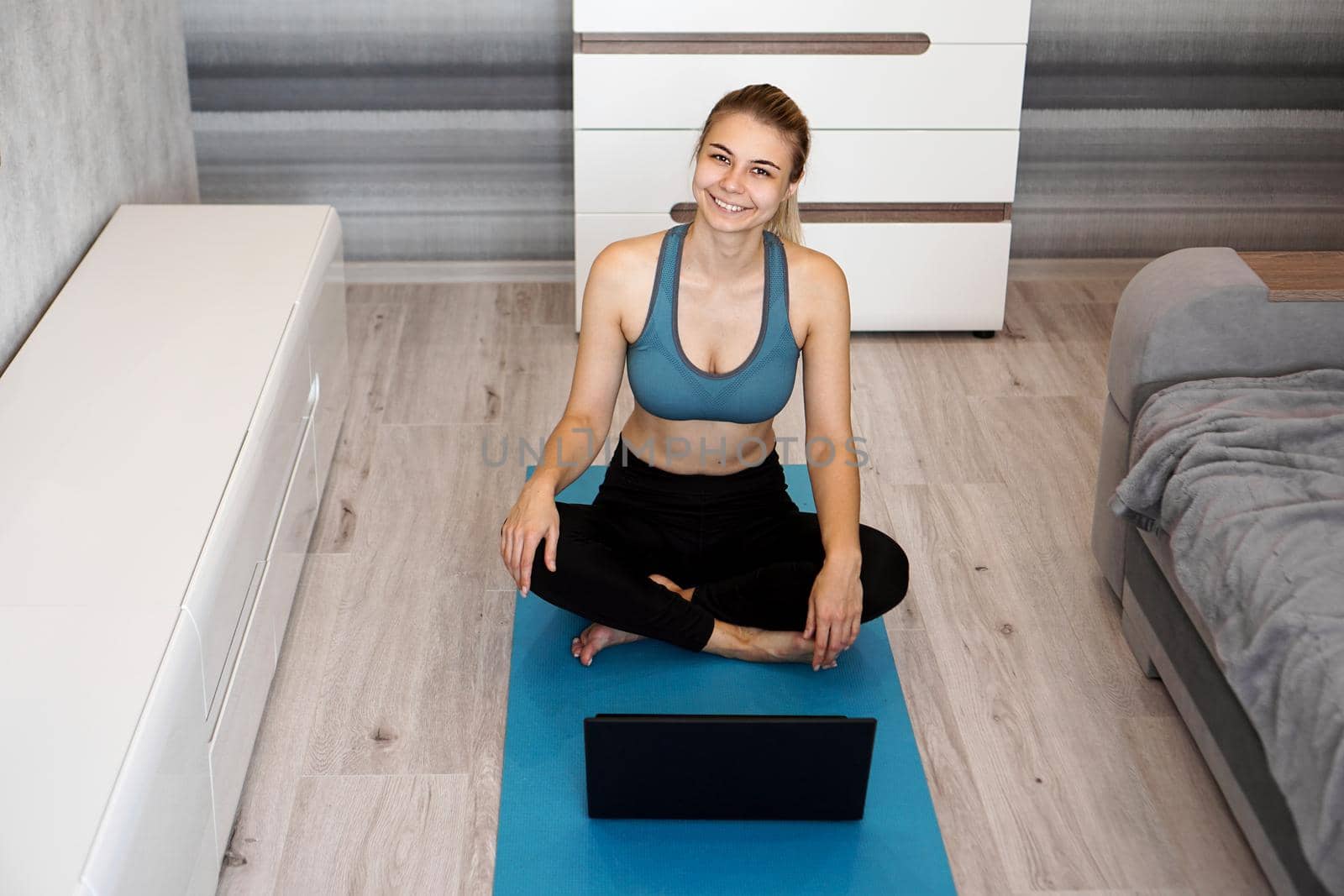 Happy young fitness woman sit on yoga mat at home near laptop by natali_brill