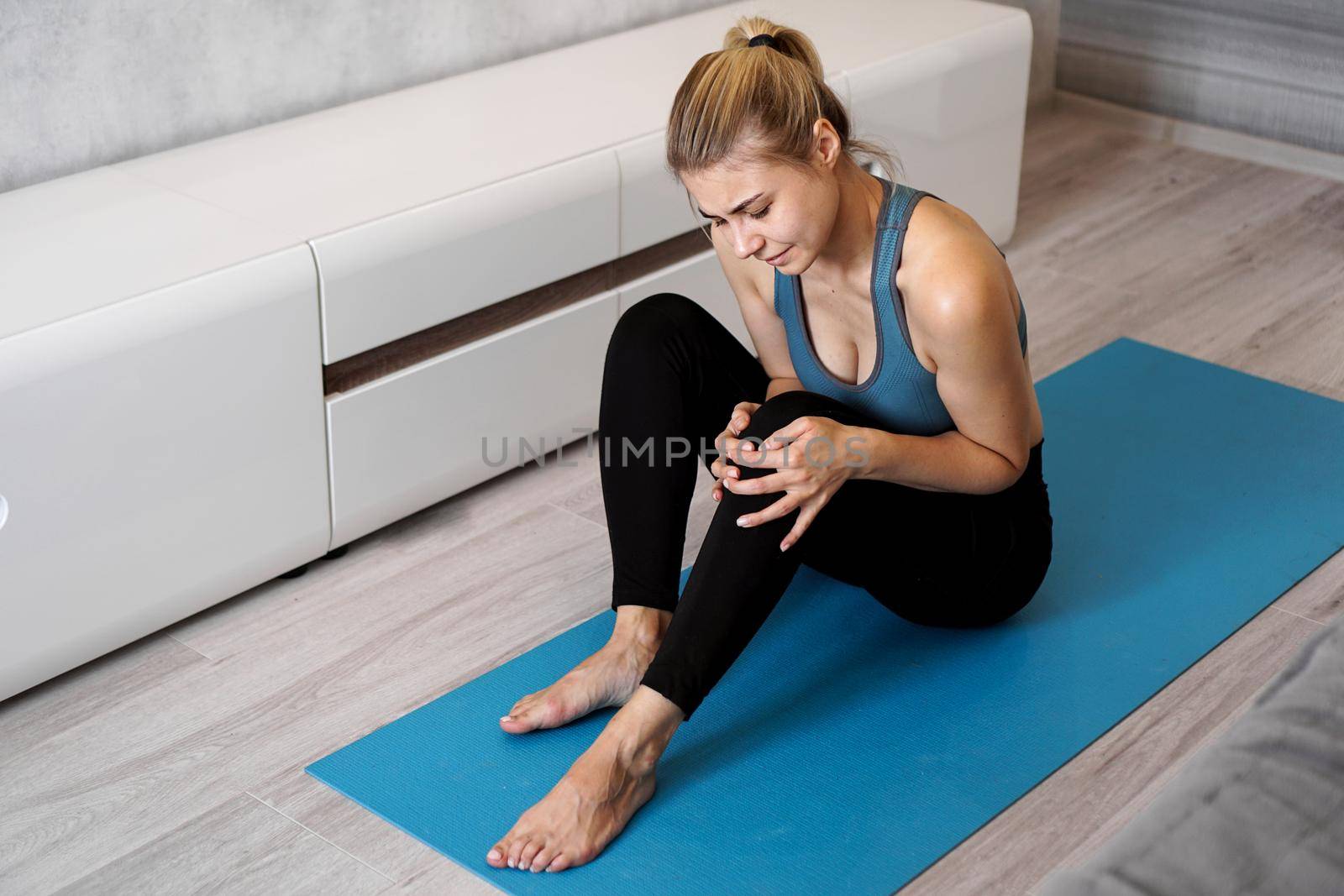 Sportswoman suffering knee ache sitting on the floor on the yoga mat in the living room