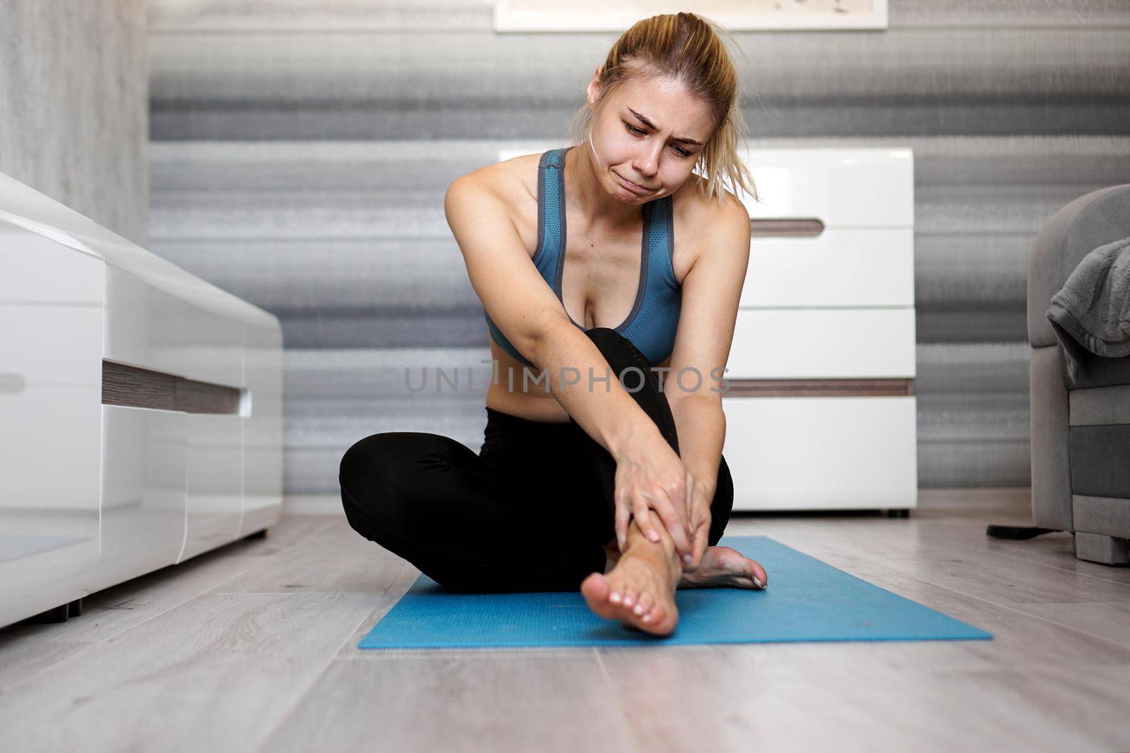 Unhappy woman sitting on the yoga mat with ankle injury, feeling pain. Health care and execise concept.