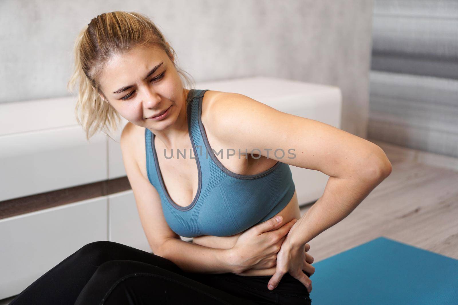 Portrait of unhappy young woman sitting on yoga mat, touching her back after training, suffering from backache, feeling pain, side view