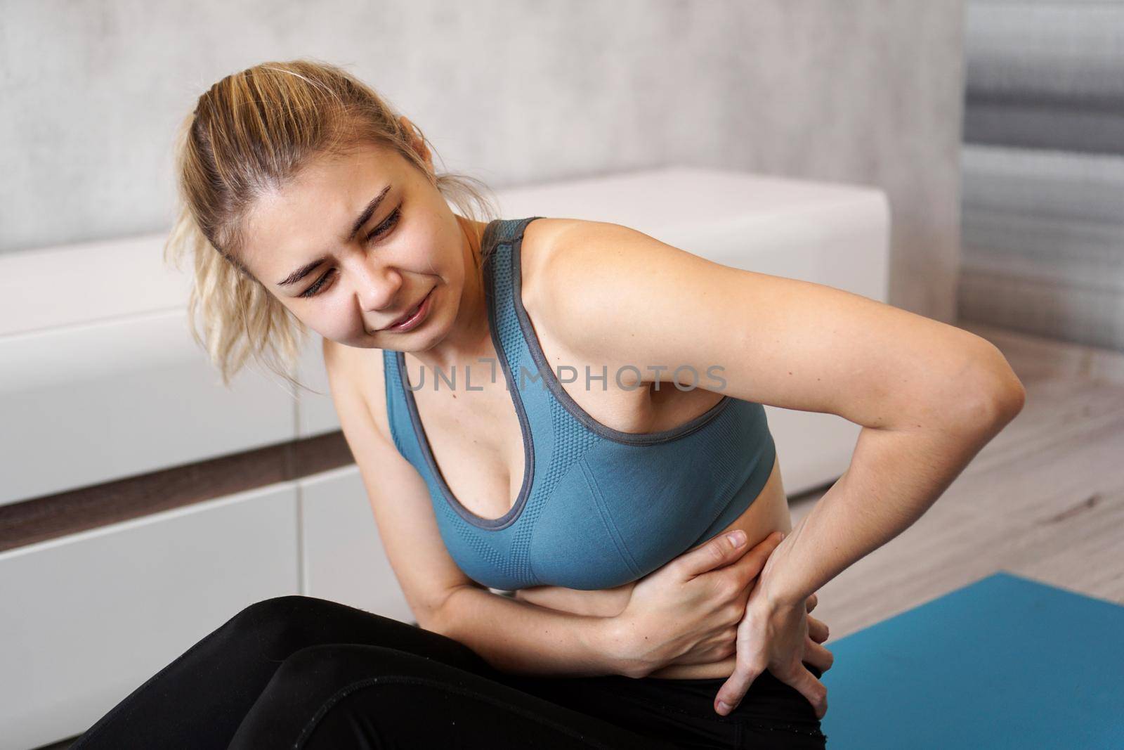 Portrait of unhappy young woman sitting on yoga mat, touching her back after training, suffering from backache, feeling pain, side view