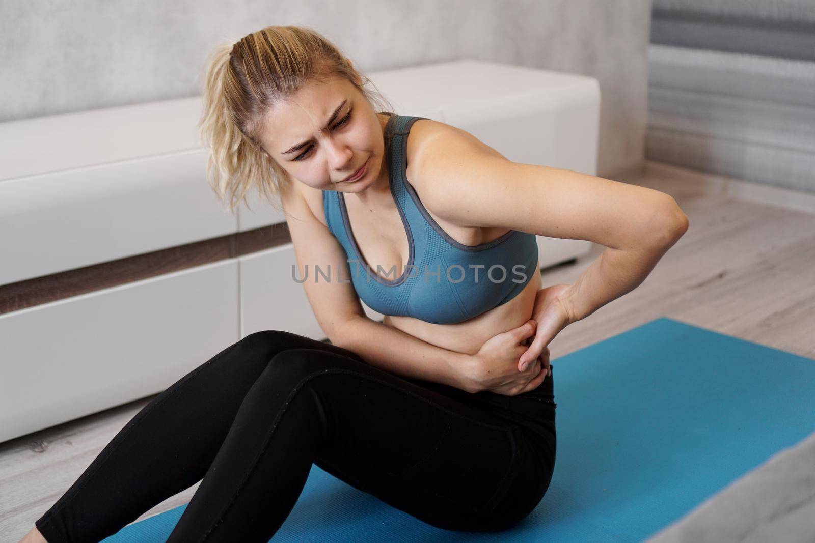 Portrait of unhappy young woman sitting on yoga mat, touching her back after training, suffering from backache, feeling pain, side view