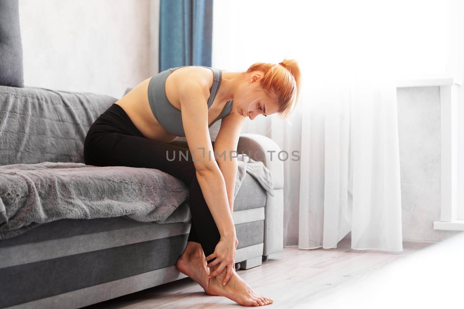 Woman sitting on sofa holds her foot injury, feeling pain. Health care concept by natali_brill