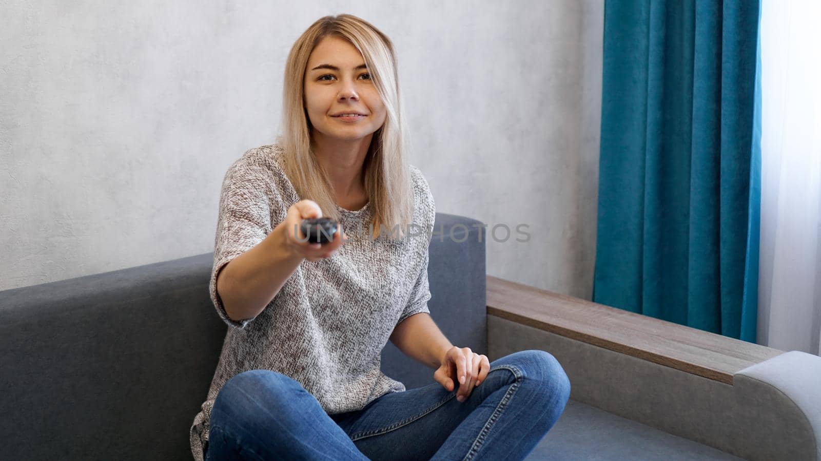 Young woman switches the television channels with the remote control. She smiles and looks at the camera