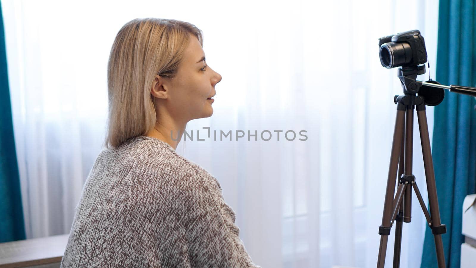 Young woman in casual clothes records herself on camera on tripod. Side view. Cheerful female blogger recording video at home