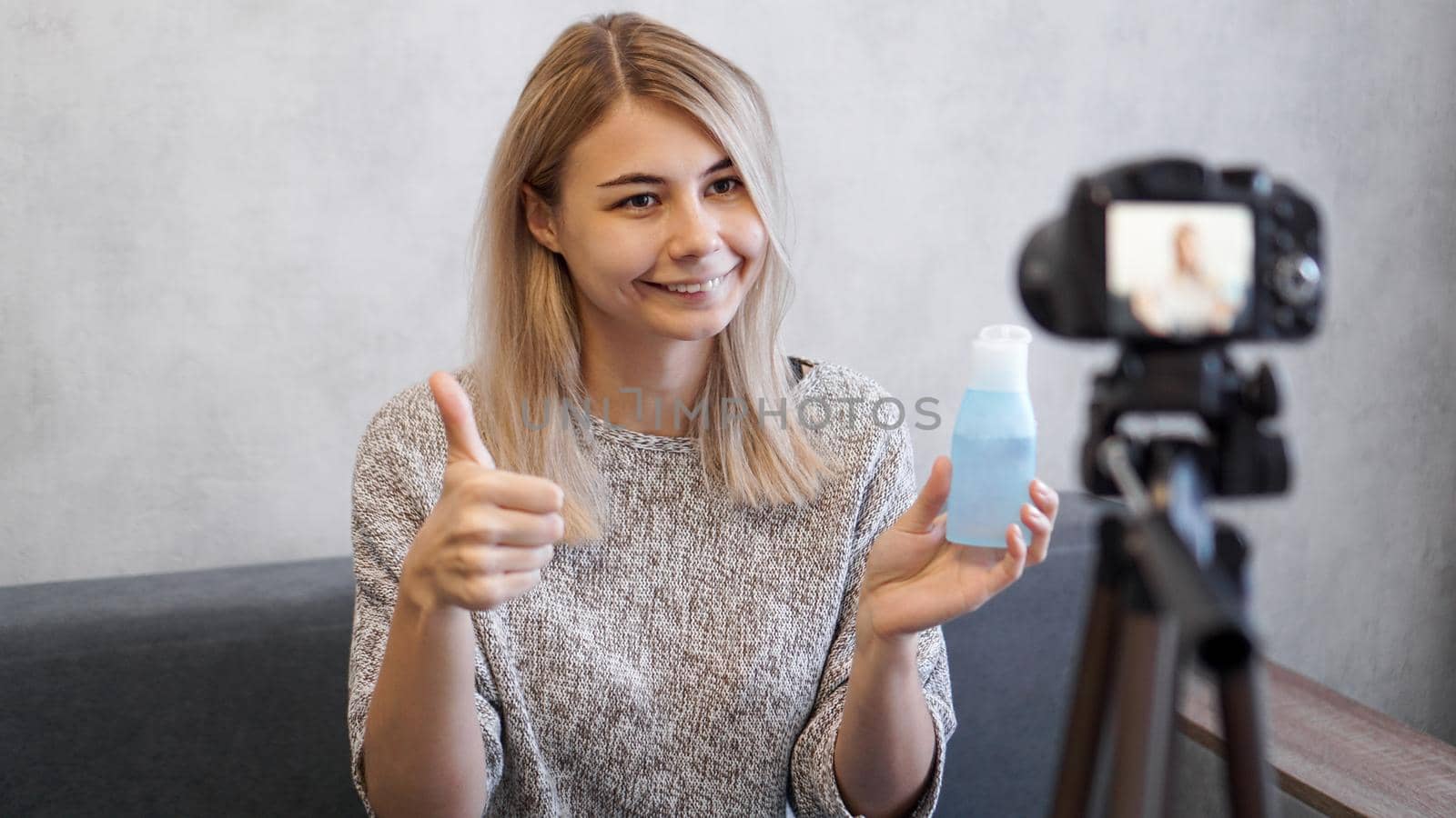 Cheerful female beauty blogger recording video at home. She shows lotion for skin and a sign like by hand