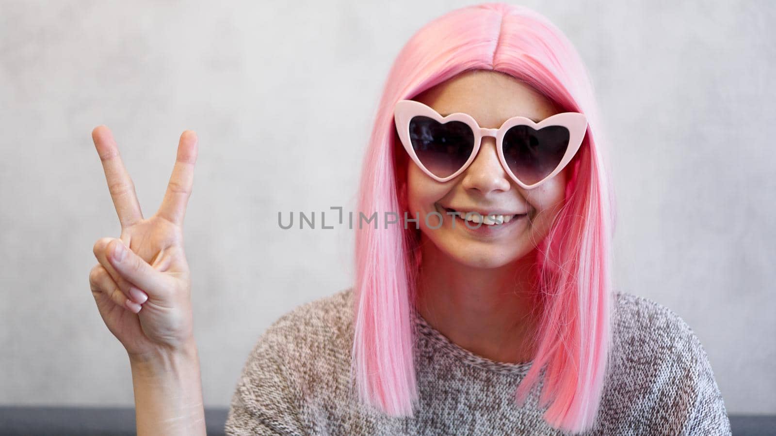 Portrait of a happy cheerful girl showing peace gesture. Woman wearing heart-shaped glasses and pink wig - positive portrait