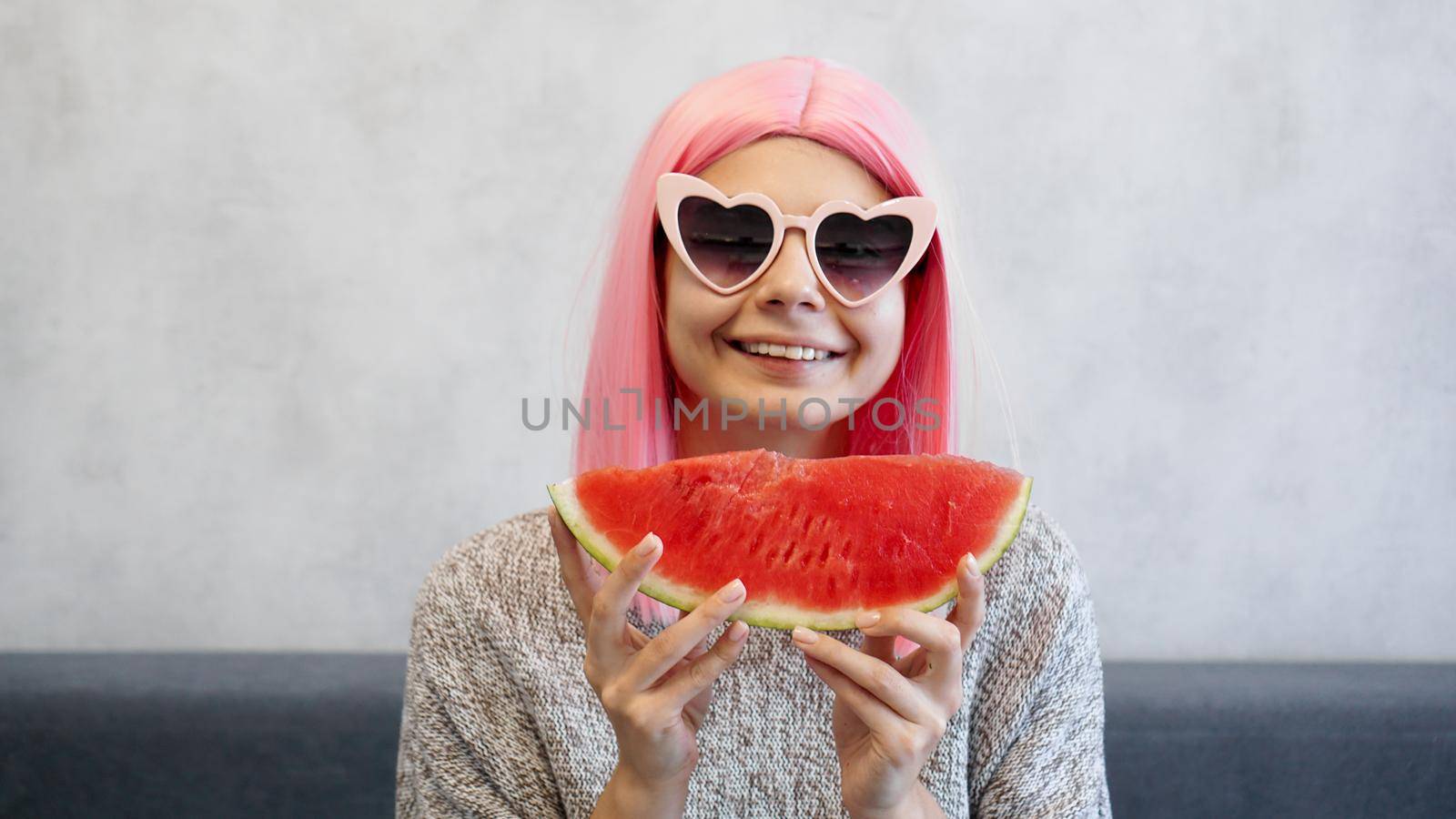 Indoor shot of young woman with piece of watermelon in hand, adorable female smiling at camera. Woman wears pink wig and heart-shaped glasses