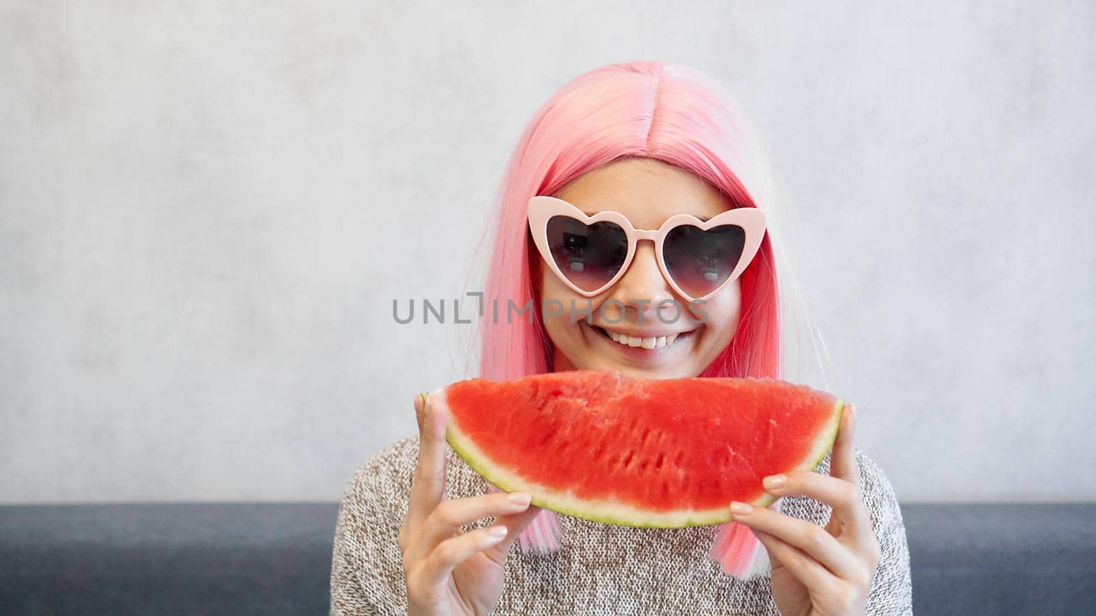 Indoor shot of young woman with piece of watermelon in hand, adorable female smiling at camera. Woman wears pink wig and heart-shaped glasses