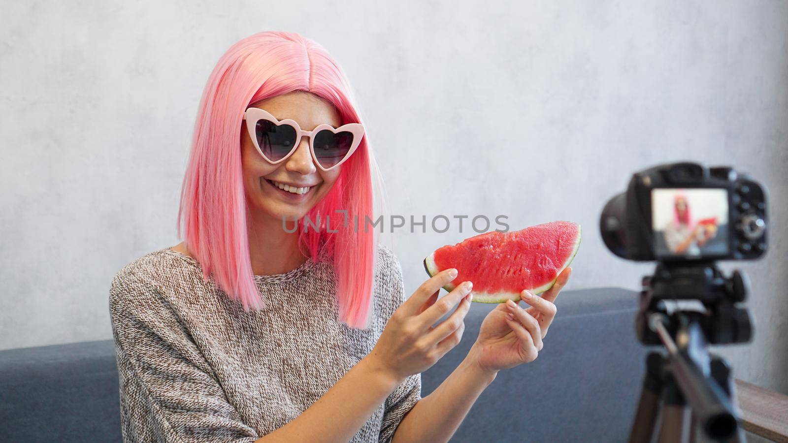 Positive blogger records a video about healthy eating and holds a watermelon in her hands. Woman wearing pink wig and heart shaped glasses