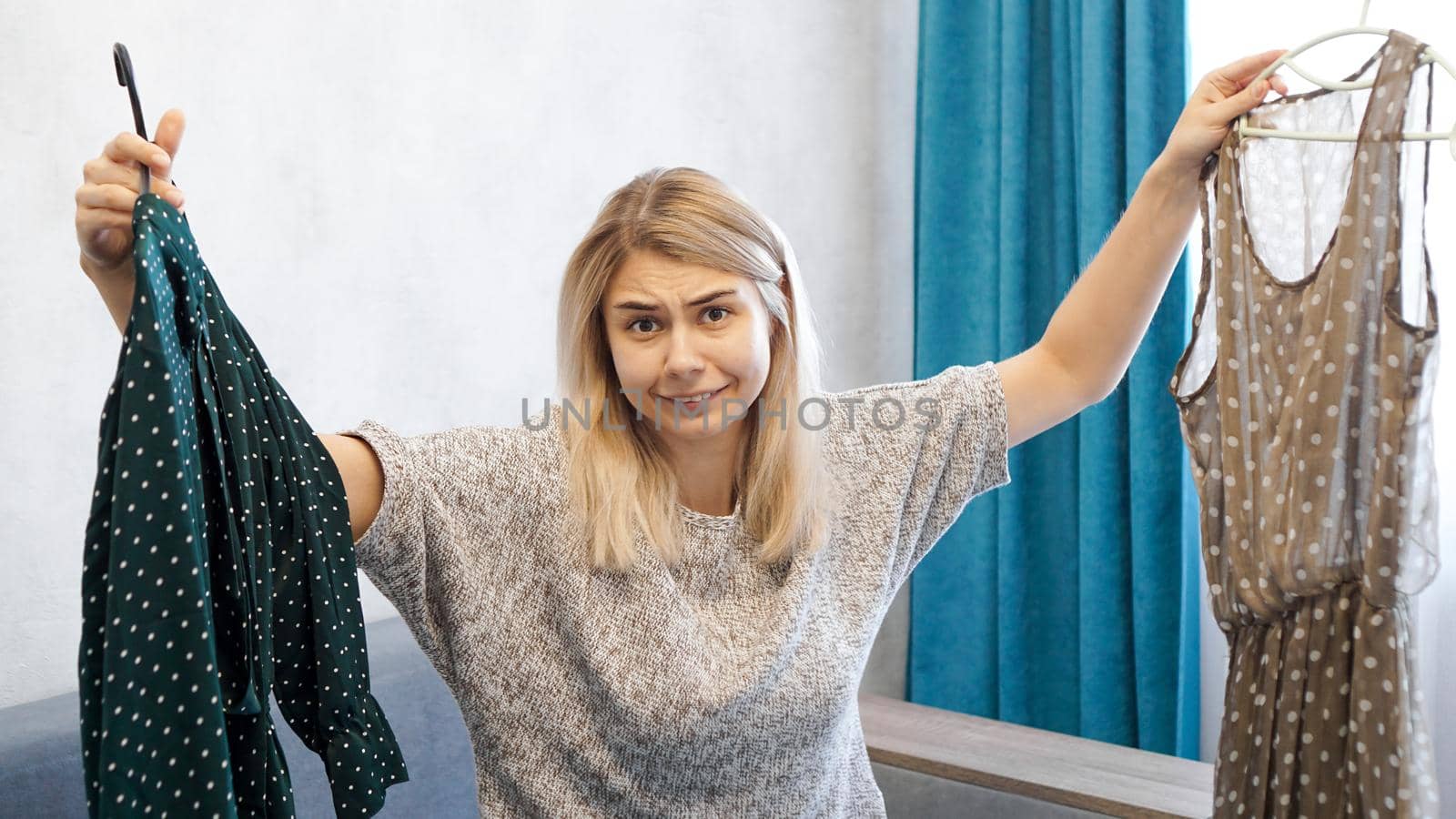 Cute woman choosing her outfit, holding two dresses on hangers. She is confused and does not know what to choose