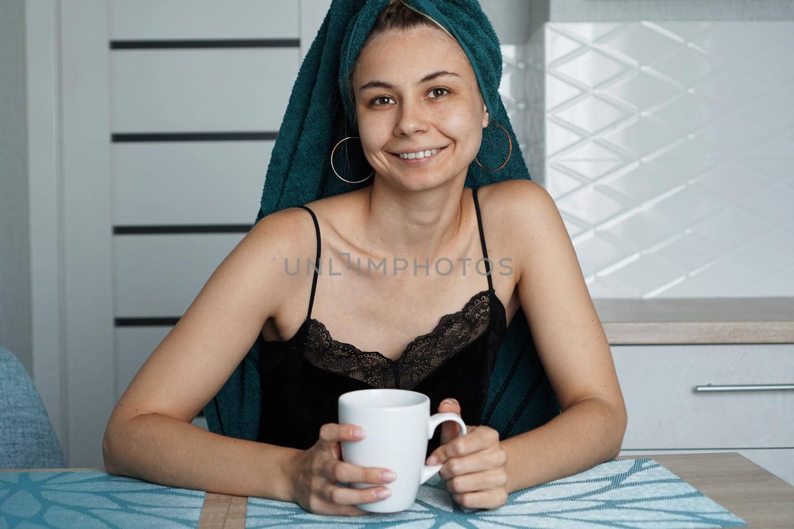 Beautiful girl in a towel on hair sits in the kitchen with a cup of coffee. Woman looking at camera and smiling
