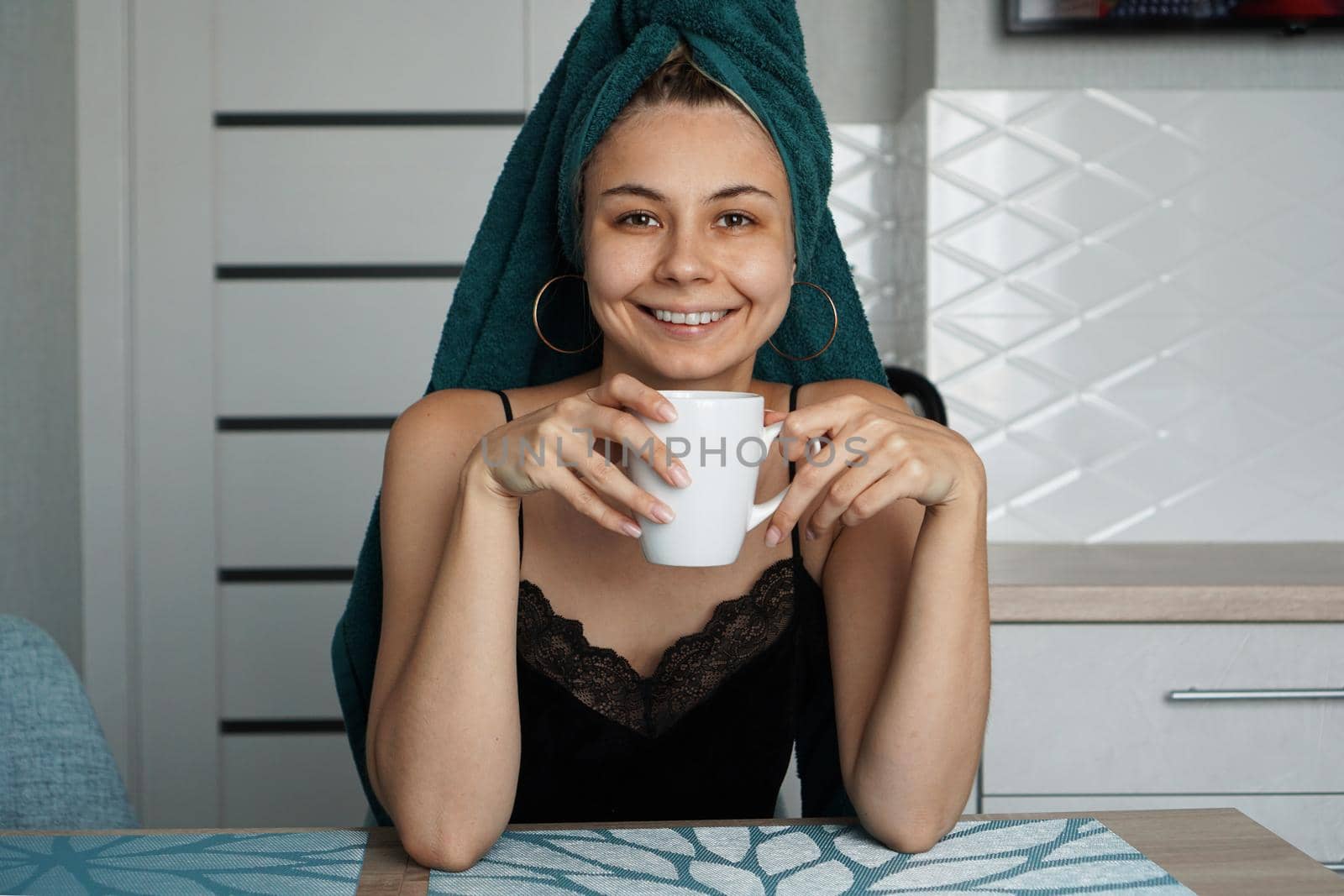 Beautiful girl in a towel on hair sits in the kitchen with a cup of coffee. Woman looking at camera and smiling