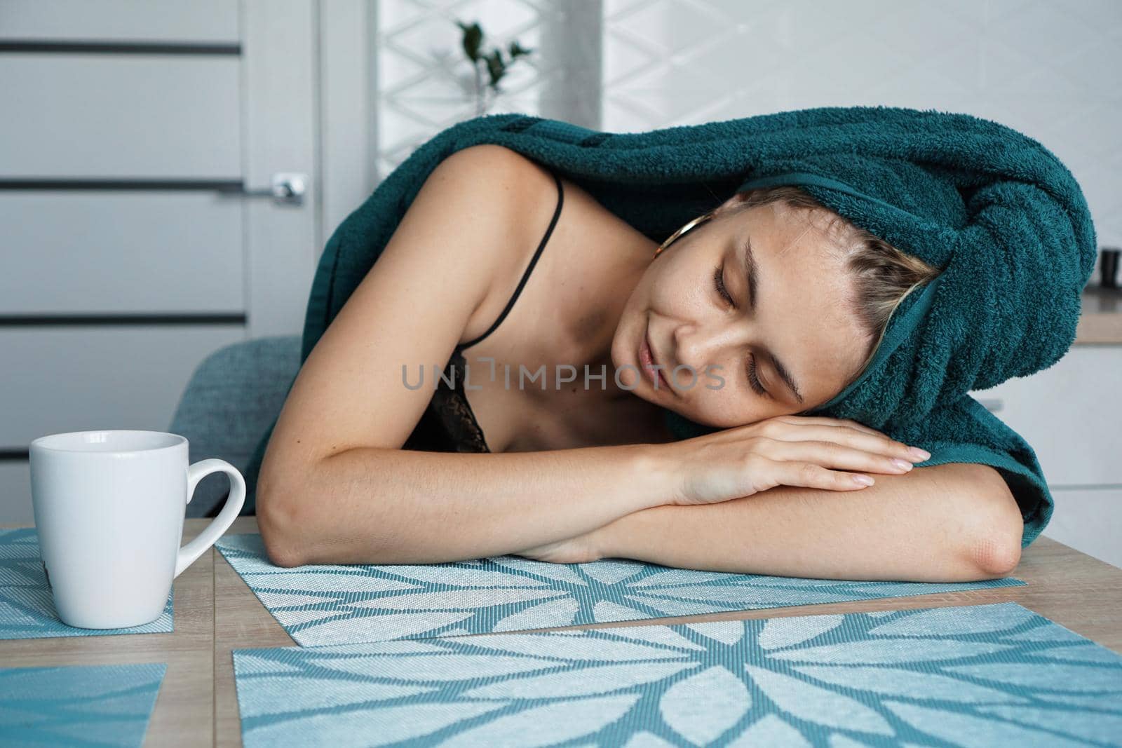 Tired woman sleeping on the table. Beautiful girl in a towel on hair sits with a cup of coffee. Lazy sleepy morning