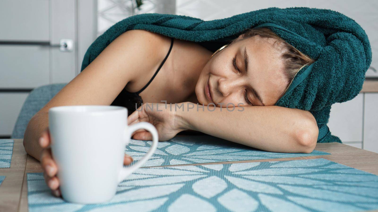 Tired woman sleeping on the table. Beautiful woman in a towel on hair. Hand reaches for the mug. Lazy sleepy morning