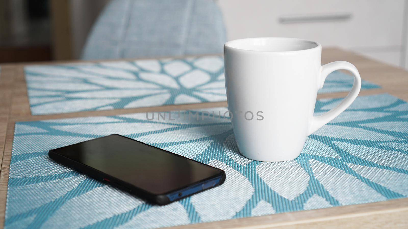 Clean white mug with handle and smart phone on blue table behind blurred kitchen background