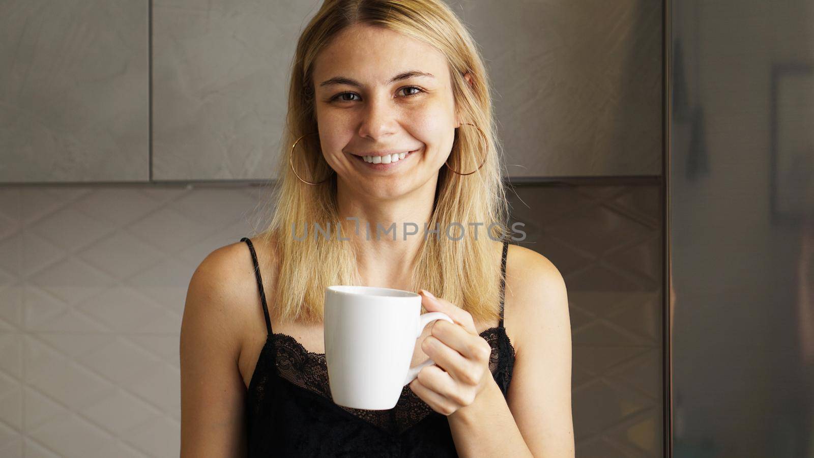 Young attractive woman with a cup of coffee smiling to camera in kitchen of modern apartment
