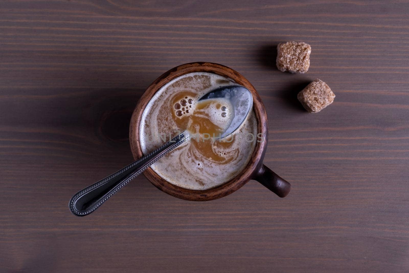 Close-up top view of hot coffee with milk in a fired earthenware mug with a small spoon and cane sugar pieces. Natural wood background.