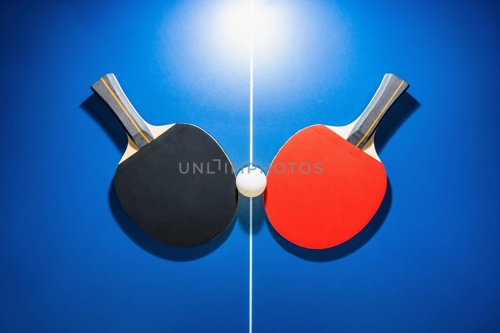 Top view black and red table tennis racket and a white ping pong ball on the blue ping pong table with a bright spotlight, Two table tennis paddle is a sports competition equipment for indoor exercise