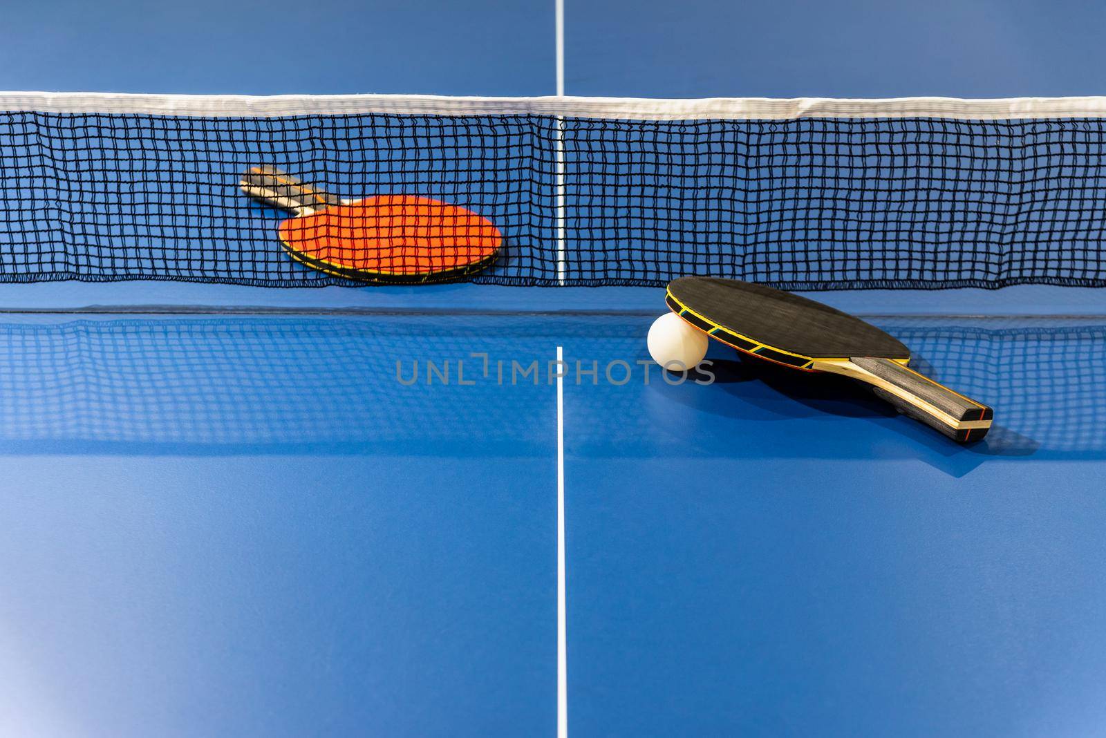 Black and red table tennis racket and a white ball on the blue ping pong table with a net, Two table tennis paddle is a sports competition equipment indoor activity and exercise for background concept