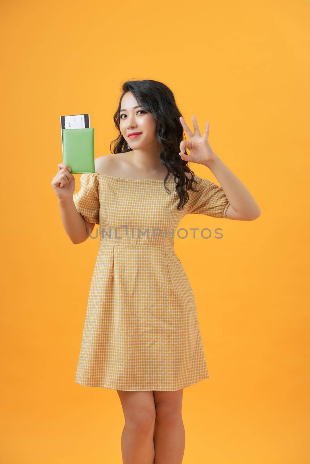Beautiful portrait young asian woman smiling gesture ok over yellow background, holding passport. by makidotvn
