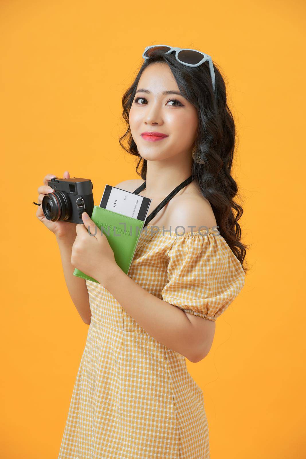 Image of happy asian young woman tourist standing isolated over yellow  background holding camera and passport with tickets.